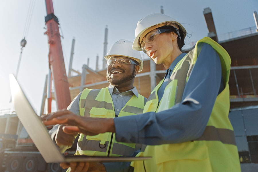 Business Insurance - View of Smiling Contractor and Engineer Standing at a Construction Jobsite and Looking at Building Plans on a Laptop