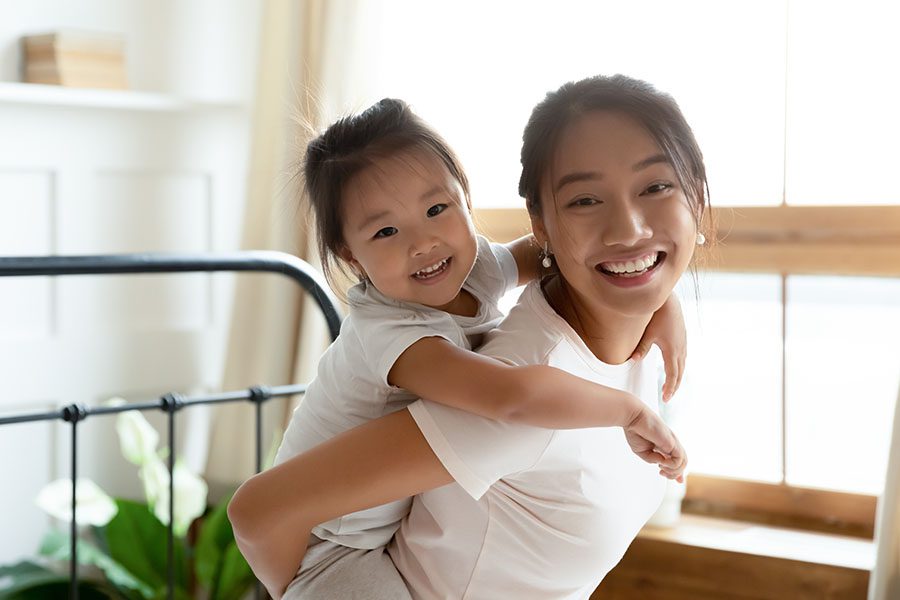 Personal Insurance - View of Smiling Mother Giving Her Happy Daughter a Piggyback Ride at Home